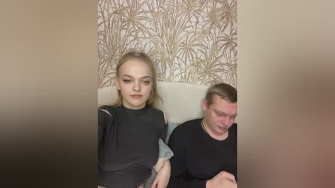 Video of a young woman with blonde hair, wearing a black top, sitting next to a man with short brown hair, both looking straight ahead. The background features a beige wallpaper with a palm tree pattern.