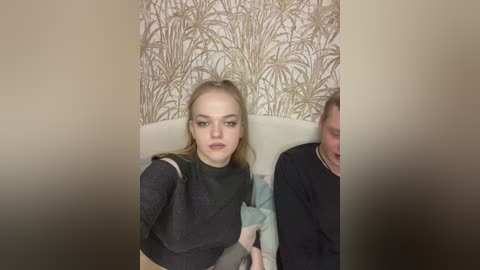 Video of a young woman with light skin and blonde hair, wearing a black dress, sitting on a white couch. Background features a beige wall with gold leaf pattern.