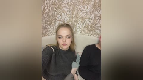 Video of a young woman with fair skin, straight blonde hair, and a neutral expression. She wears a black lace top, sitting in a beige chair against a wallpaper with gold palm leaf pattern.