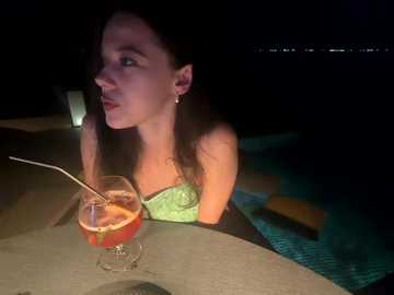 Video of a fair-skinned woman with long dark hair, wearing a strapless green top, holding a drink with a straw near a pool at night.
