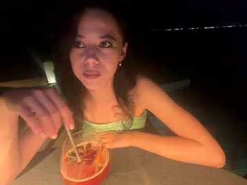 A dimly lit video of a young woman with long dark hair, wearing a green top, holding a glass of red drink with a straw, in a dark, possibly night-time outdoor setting.