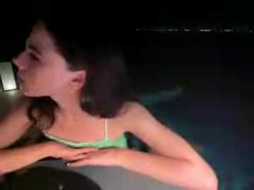 Video of a young woman with shoulder-length brown hair, wearing a green spaghetti-strap top, lying on her side at night. She has a contemplative expression. Background shows dark night sky and faint lights.