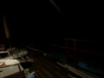 A dimly lit video captures a dark, shadowy indoor scene with a cluttered table and scattered objects on the left, a red shelf on the right, and a pool visible through a window.