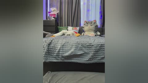 Video of a cozy bedroom with a grey knitted blanket on a bed, featuring a plush cat toy and a stuffed animal dog, grey curtains, and a nightstand with a pink flower decoration.