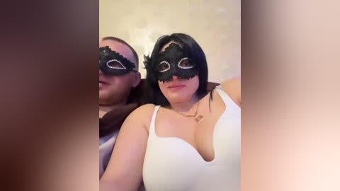 A video of a woman with long black hair wearing a revealing white top and black masquerade mask, standing beside a man in a brown shirt, both indoors.