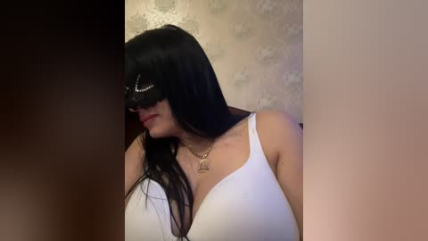 Video of a woman with long black hair, wearing a white bra and a gold necklace, standing against a floral wallpaper.