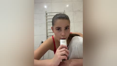 Video of a young woman with light skin, dark hair in a ponytail, wearing a red top, holding a white bottle to her mouth in a tiled bathroom.