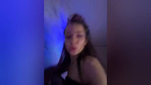 Video of a young woman with long dark hair, wearing a black top, against a blue-lit wall, mouth open, blurry background.