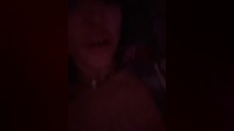 A dimly lit, blurred video captures a close-up of a woman's face and shoulders, partially obscured by shadow. Her skin appears pale, and she has long, dark hair. The background is indistinguishable due to the low light.