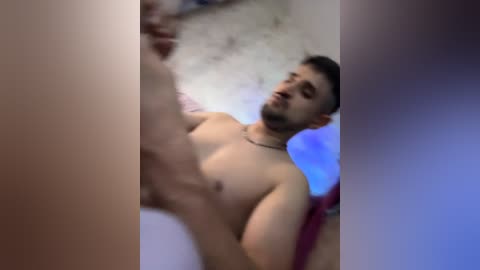 A blurred video of a topless, muscular, bearded man with short dark hair, wearing a necklace, standing in a dimly lit room with peeling wallpaper.