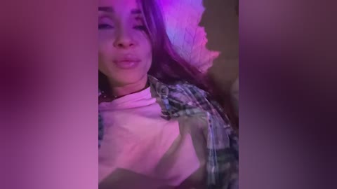 A video of a young woman with light brown skin, wearing a white shirt and plaid jacket, her hair partially hidden by a pink scarf. The background is blurred, suggesting a dimly lit indoor setting.