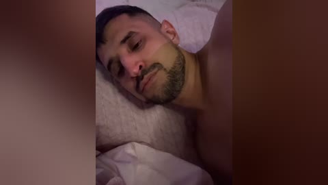 Video of a sleeping man with short dark hair and a trimmed beard, lying on his side under white sheets, eyes closed, looking peaceful.