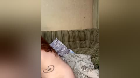 Video of a young woman with red hair, partially visible, lying on a striped, beige sofa, covered in a patterned blanket. The room has a beige wall with a subtle texture.
