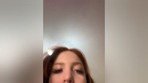 A blurry video of a young woman with long, reddish-brown hair and fair skin, wearing mascara and eyeliner, with closed eyes, standing against a plain, light-colored background.