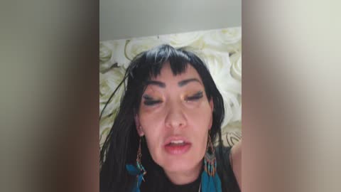 Video of an Asian woman with long black hair and bangs, wearing a black sleeveless top, lying on a bed with white floral sheets, eyes closed, expression serene.