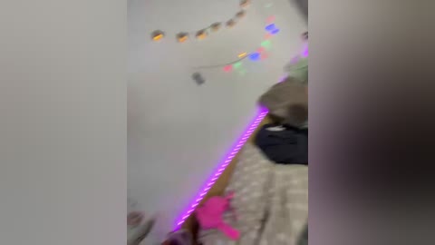 A blurred video of a room with a white wall, colorful string lights, and a strip of purple LED lights along the floor. The background is out of focus, making the room indistinct.