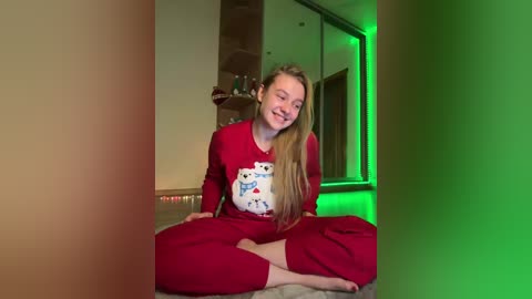 Video of a smiling young woman with long blonde hair, wearing red pajamas with a cartoon character design, sitting cross-legged on a carpet in a dimly lit room with green LED lighting.