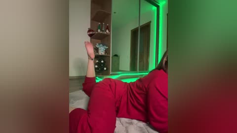Video of a woman lying on her stomach in a bedroom, wearing red pajamas, with her legs bent and feet raised, under a green light, beige walls, wooden shelves, and a mirrored closet in the background.