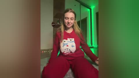 A video of a young woman with blonde hair, wearing red pajamas featuring a cartoon bear, sitting on a bed in a dimly lit bedroom with green neon lights.