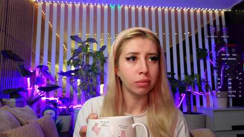 Video of a young, fair-skinned blonde woman with long hair, wearing a white t-shirt, holding a cup, indoors with vertical blinds, potted plants, and colorful string lights in the background.