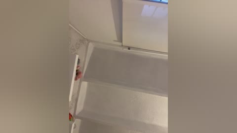 A video of a small, modern bathroom with white walls, a white bathtub, and a white toilet. The ceiling has a recessed light fixture. A pink toothbrush and a red object are placed on the white countertop.