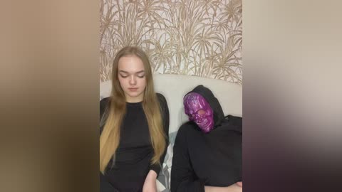 Video of two young women, one in a black dress with long blonde hair, the other in a purple mask, sitting on a sofa against a wallpaper with abstract patterns.