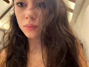 Video of a young woman with long, dark, wavy hair partially covering her face, wearing a brown top. She has fair skin and blue eyes, and is indoors with a blurred background.