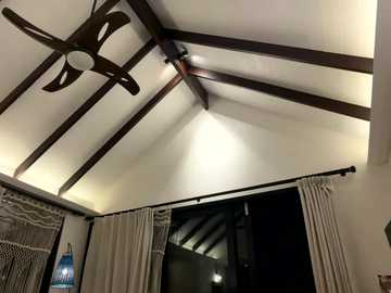 Video of a dimly lit, cozy bedroom with exposed wooden ceiling beams, beige curtains, a large mirror, and a ceiling fan.