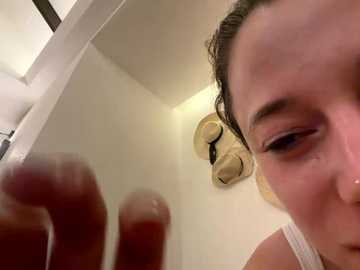 A close-up video of a woman with wet hair, wearing a white tank top, reaching toward a mirror. The background shows a beige ceiling fan and part of a room with white walls.