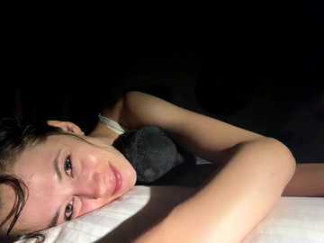 Video of a young Asian woman lying on a bed, illuminated by a dim light. She has light skin, dark hair, and is wearing a black bra. The background is dark and indistinct.