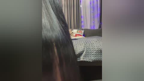 A video of a cozy bedroom with a dark-haired person in the foreground, a gray bed with a white and red pillow, and string lights in the background.