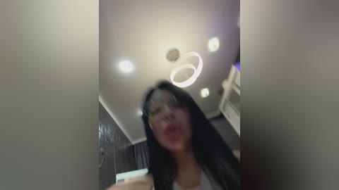 A video of a woman with long black hair, wearing a white tank top, standing in a dimly lit room with a circular light fixture.