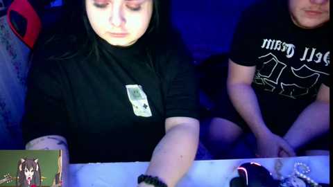 Video of two women, one with long black hair, wearing a black t-shirt, seated at a table in a dimly lit room.