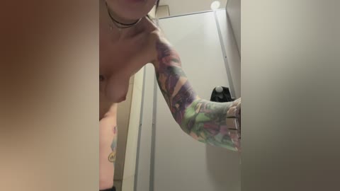 A video of a topless woman with colorful arm tattoos, standing in a bathroom, leaning against a white shower stall, wearing a choker necklace.