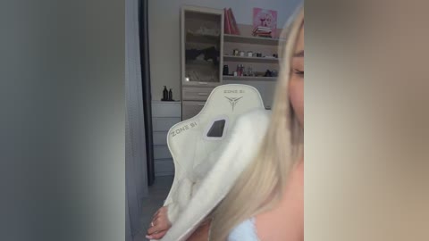 Video of a blonde woman in a white tank top, holding a white Converse shoe, taken in a modern, minimalist bedroom with light wood shelves and a pink storage unit.