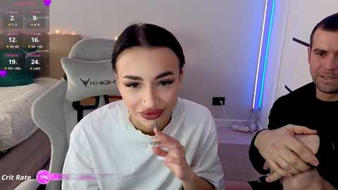 Video of a young woman with dark hair and fair skin, wearing a white t-shirt, sitting on a grey chair. A man with short hair and a beard is seated beside her. The background features a modern room with a bed, dim lighting, and a gaming chair.