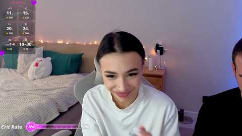 Video of a young woman with dark hair, smiling, sitting on a grey chair in a cozy bedroom with a bed, teal pillows, and a stuffed bear.