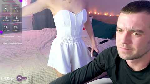 Video of a white woman in a white strapless dress on a bed, holding a remote, with a white man in a black shirt watching, both in a bedroom with candles and a quilted bedspread.