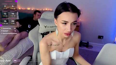 A video of a slender, dark-haired woman with a small tattoo on her collarbone, wearing a white strapless dress, sitting on a gray gaming chair in a dimly lit room with a bed and a man in the background.