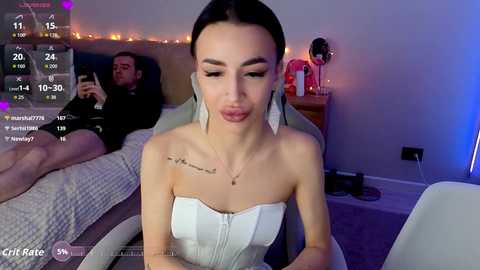 Video of a woman in a strapless white dress with long dark hair, sitting in a dimly lit room with a man on a bed, streaming on \"Chaturbate.\