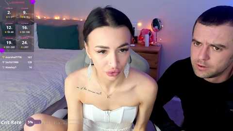 Video of a young, fair-skinned woman with dark hair, wearing a strapless white dress, sitting on a bed, making a kissing face. A man with short hair, wearing a black shirt, is behind her.
