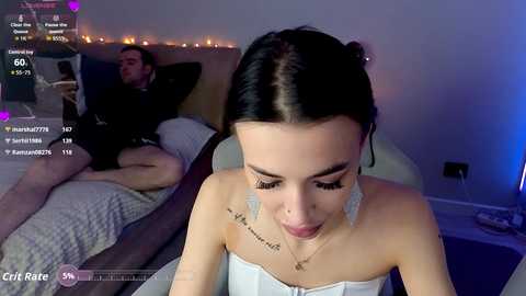 A video of a young woman with fair skin, black hair, and silver earrings, wearing a strapless white dress, sitting on a bed with a man in black, watching a video on a phone.