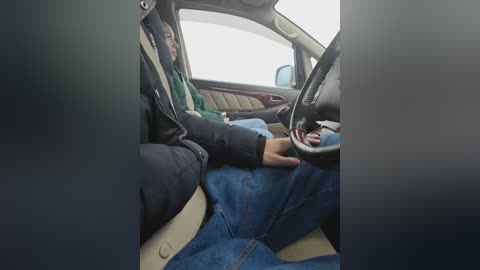 A video of a person in a car, wearing a black jacket, green shirt, and blue jeans, holding the steering wheel, with beige seats and dark dashboard visible.