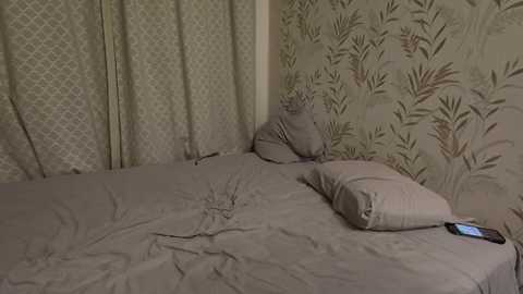 Video of a small, cluttered bedroom with a messy bed, gray bedding, a pillow, and a smartphone on the mattress. The wallpaper features a green floral pattern, and the curtains are light with a diamond design.