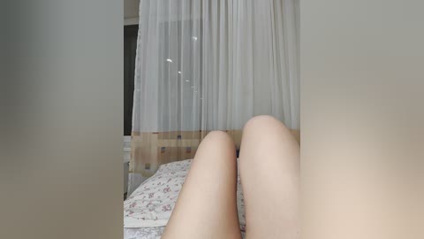 Video of a woman's bare legs, lying on a floral-patterned bed, with sheer white curtains in the background.