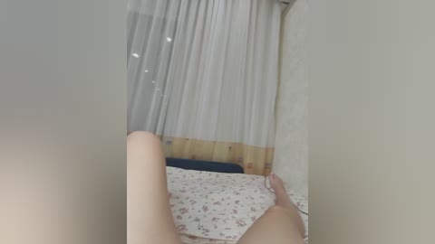 Video of a light-skinned person lying on a bed with white sheets, legs slightly apart, viewed from above. The background features a window with white curtains and wooden trim.