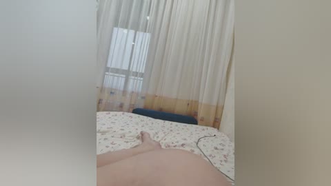 A video of a dimly lit bedroom with sheer white curtains, a bed covered in floral sheets, and a hand resting on the bed.