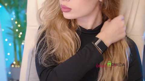 Video of a young woman with long, wavy blonde hair, wearing a black top, standing in front of a beige wall with a blurred green plant and string lights in the background.