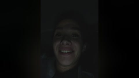 A dimly lit video of a young boy with light skin and curly hair, smiling broadly. His face is slightly obscured by a shadow, adding a mysterious, playful atmosphere.