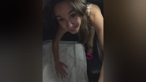 A video of a young woman with long, dark hair and fair skin, leaning over a white towel on a bed. She wears a sleeveless top and pink bracelet. The background is dimly lit.